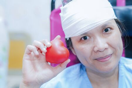 Asian middle-aged lady woman patient  holding red heart in her hand on bed in nursing hospital ward : healthy strong medical concept.の写真素材