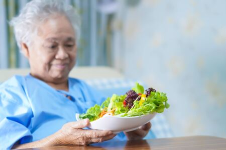 Asian senior or elderly old lady woman patient eating breakfast vegetable healthy food with hope and happy while sitting and hungry on bed in hospital.の写真素材