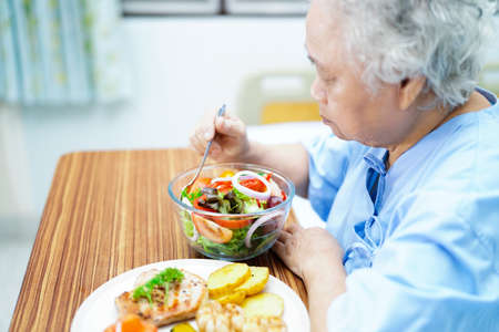 Asian senior or elderly old lady woman patient eating breakfast vegetable healthy food with hope and happy while sitting and hungry on bed in hospital.の写真素材