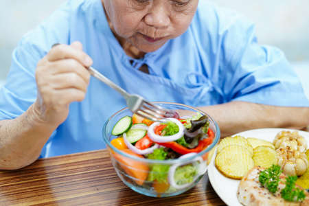 Asian senior or elderly old lady woman patient eating breakfast vegetable healthy food with hope and happy while sitting and hungry on bed in hospital.の写真素材
