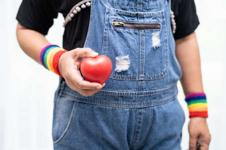 Asian lady wearing rainbow flag wristbands and hold red heart, symbol of LGBT pride month celebrate...