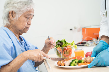 Asian senior or elderly old lady woman patient eating breakfast and vegetable healthy food with hope and happy while sitting and hungry on bed in hospital.の写真素材