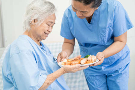 Asian senior or elderly old lady woman patient eating breakfast and vegetable healthy food with hope and happy while sitting and hungry on bed in hospital.の写真素材