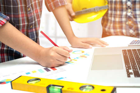 Asian engineer and partner team learn and talk with house plan project blueprint, graph and yellow helmet and engineering construction tools on table work.の写真素材