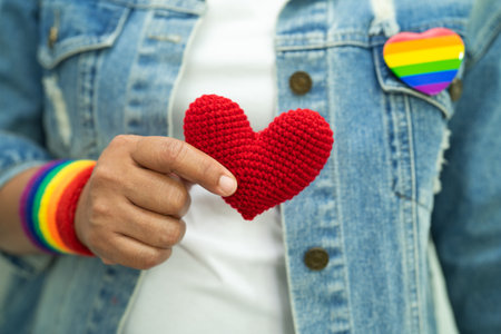 Asian lady wearing rainbow flag wristbands and hold red heart, symbol of LGBT pride month celebrate...