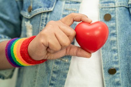 Asian lady wearing rainbow flag wristbands and hold red heart, symbol of LGBT pride month celebrate annual in June social of gay, lesbian, bisexual, transgender, human rights.の写真素材
