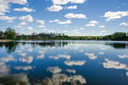 Cloudy blue sky reflects into the lake with trees on the horizon.の写真素材