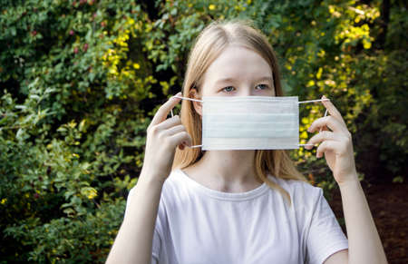 A young teen girl on the street puts on a medical mask. Portrait of a child with light hair in a...