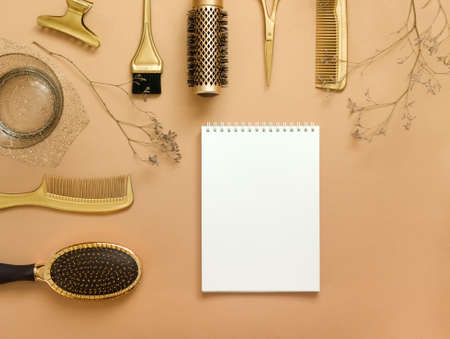 The background is beige with hairdressing tools, an empty white notebook, and dried flowers. Gold hair accessories, pattern with copy space.の写真素材