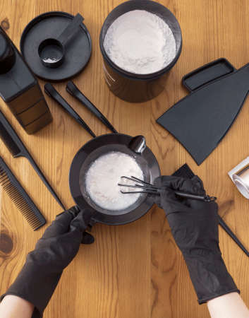 Womens hands in black gloves hold a bowl of paint and prepare tools for hair dyeing. Accessories on a wooden table, top view. Coloring and bleaching of hair at home or in a hair salonの写真素材