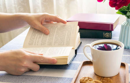 Reading a book at a table by the window. Book in hand, morning coffee at home or in a cafe, cozy...