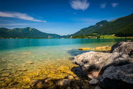 Rocks in front of the Sea in Austria called Wolfgangsee with clear water which turquoise is blue and mountains in the background and clouds in the skyの写真素材