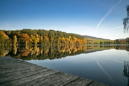 Footbridge on a lake in the autumn with orange and yellow trees and reflection from the forest on the water in the bavarian forestの写真素材