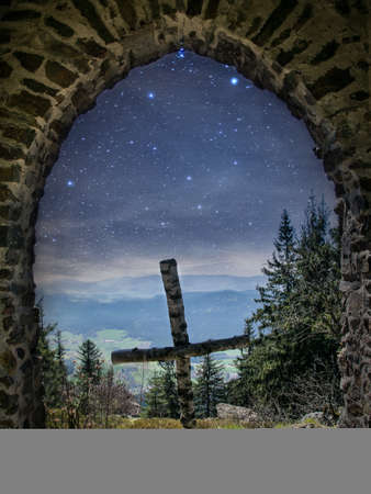 Cross on a mountain with view through a brick arch in the valley with stars on the sky in the bavarian forestの写真素材