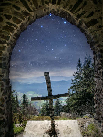 Cross on a mountain with view through a brick arch in the valley with stars on the sky in the bavarian forestの写真素材