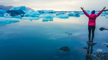 A girl in pink jacket standing on the rock in a shallow water. She spreads her arms up and wide. Glacier lagoon full of drifting ice bergs. Global warming causing melting of glaciers. Cold tones.の写真素材