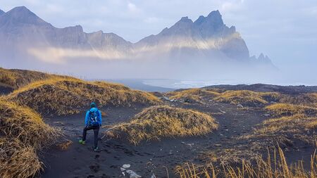 A young man wearing blue jackets stands between black sand dunes and stares at the tall mountains....