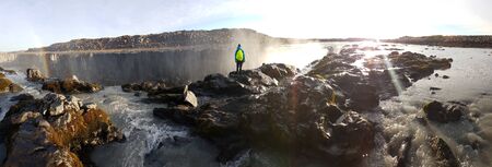 A young man standing on top of a waterfall. He is wearing a blue jacket and  neon yellow backpack....