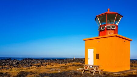 A cute little, orange lighthouse placed on the shore of a steep cliff. In front of the lighthouse...