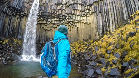 A young man wearing hiking backpack, standing in front of a waterfall, takes a selfie. Rocks on the...