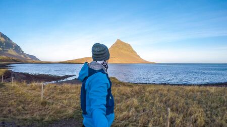 A young man wearing blue jacket and a beanie walks around famous Kirkjufell mountain in Iceland. Sea surrounding the hat like mountain. Beautiful and clear weather. Man taking a selfieの写真素材