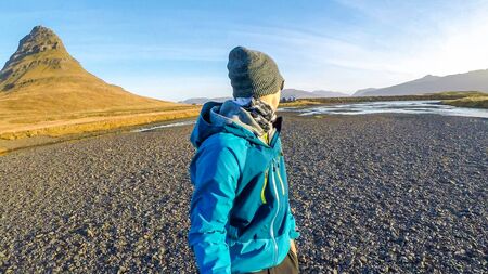 A young man wearing blue jacket and a beanie walks around famous Kirkjufell mountain in Iceland. Sea surrounding the hat like mountain. Beautiful and clear weather. Man taking a selfieの写真素材