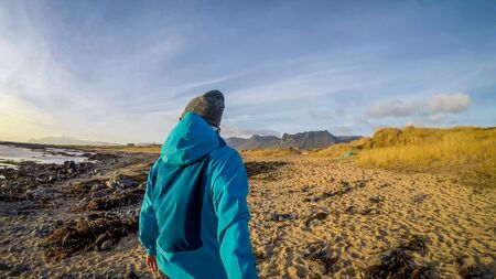 A young man wearing blue jacket and a beanie walks around flat grassland, with sea in front of him. He is holding a selfie stick, and taking pictures. Beautiful and sunny day. Exploring the nature.の写真素材