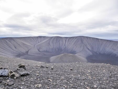 A view from the top into the volcano crater. Volcanic landscape. Ground is covered with volcanic dust and small pebbles and rocks. Overcast, with sun trying to get through the thick clouds.の写真素材