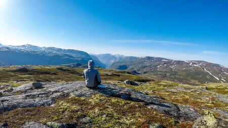 A young man wearing a hoodie sit at the ground and enjoys the endless mountain view of Eidfjord. He...