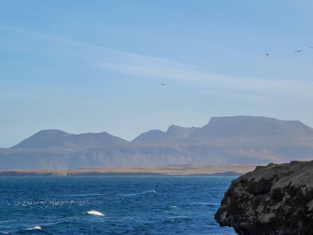 A wavy shore of a fjord, with tall mountains as a backdrop. In the front on the side is a sharp rock...