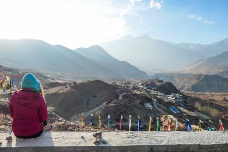 A girl wearing pink jacket sitting on a concrete wall on a top of a mountain and enjoying the misty Himalaya range spreading in front of her. The sun light is nicely marked. Freedom and achievement.の写真素材