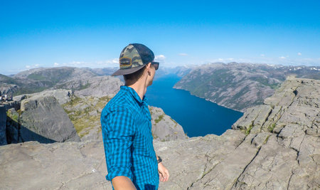 A young man in a hiking outfit walks along the edge of a steep cliff , with a selfie stick. He walks on a barren rocky cliffs. In the back is Lysefjorden, Norway. Bright and sunny day.の写真素材
