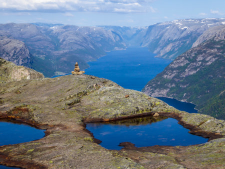 A stone-man build at the edge of a steep cliff  with the view on Lysefjorden. Stone-man is guarding the surrounding area. Picturesque view on he fjord. Few puddles on top of the cliff,の写真素材