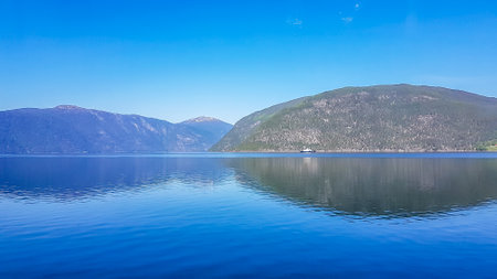 A view on the Songefjorden (King of the Fjords) from the water level. It is the deepest fjord in Norway. Tall, lush green mountains surrounding the fjord. Calm surface of the water. Clear blue sky.の写真素材