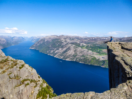 A man wearing blue shirt sits at the edge of a steep cliff of Preikestolen with his legs hanging down. A view on Lysefjorden. Fjord goes far inland. Man enjoys the view, feels free and happy.の写真素材