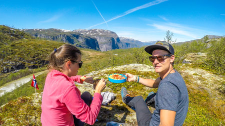 A couple having a meal in the tall mountains. Girl is eating the noodles with a fork. Boy is holding his portion in one hand, taking selfie with another hand. Camping in Norwegian wilderness.の写真素材