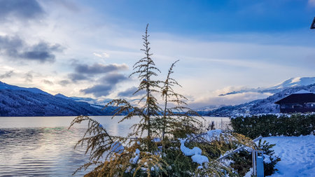A beautiful view on a MillstÃ¤tter lake in Austria. The lake is surrounded by Alps. Mountains are covered with snow. Soft colors of the sunset. Blue sky. In the middle there is a tree.の写真素材