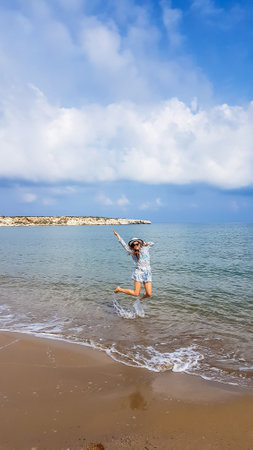 A young woman in blue dress and hat jumps over the waves in the sea, Lara Beach, Cyprus. Turquoise color of the water.  Happiness and freedom. Solo traveling girl. Clear but cloudy weather.の写真素材