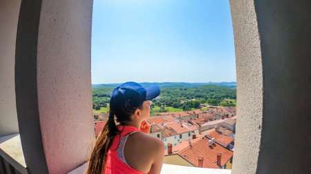 A girl looking through the window of bell tower on the harbor. Rows of docks waiting for the boats and yachts to anchor there. Lots of houses surrounding the port. In the back there are few islandsの写真素材