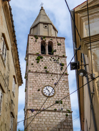 Bell tower located in the middle of an archival city. Tower is towering high above other buildings. Captured from the bottom. All the buildings are coloured beige. Tower overgrown with plants, flowersの写真素材