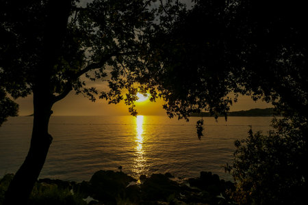 Romantic sunset by a beach. The sun sets over the horizon. The sun beams reflecting in the calm sea waters. There is an island on the side.Trees growing on the shore. The sky turns yellow and orangeの写真素材