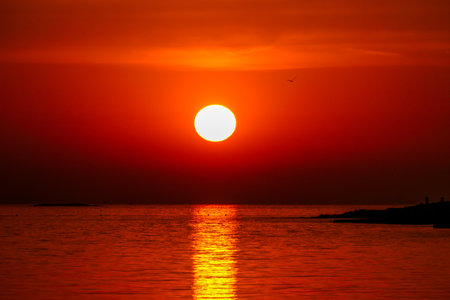 Romantic sunset by a beach. The sun sets over the horizon. The sun beams reflecting in the calm sea waters. There is an island on the side. Few birds flying around. The sky turns yellow and orangeの写真素材