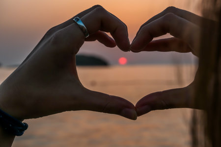 A heart formed from hands, and framing the setting sun in it. The sun sets over the horizon. The sun beams reflecting in the calm sea waters. The sky turns yellow and orange. An island on the sideの写真素材