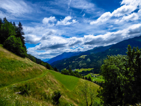 A stunning view on Alpine valley. Hiking trails in the high mountain. Green grass and bushes cover the slopes. A remote view on small settlement in the bottom of the valley. Spring in the mountains.の写真素材