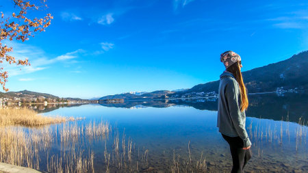 Girl wearing a beanie walks along the lakeside. Calm surface of the lake reflects the mountains and...