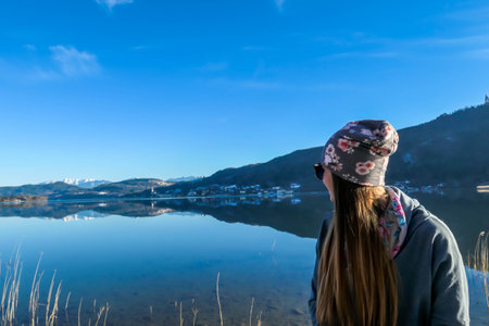 Girl wearing a beanie poses with the lake view behind her. Calm surface of the lake reflects the mountains and the sky. Girl is smiling, enjoying her time and beautiful weather. Happy autumn moment.の写真素材