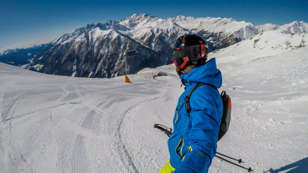 A young skier takes a selfie while skiing down a perfectly groomed slope. Man is wearing helm for...