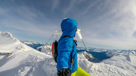 A man in skiing outfit standing on the top of a mountain peak with a snow covered cross behind him....