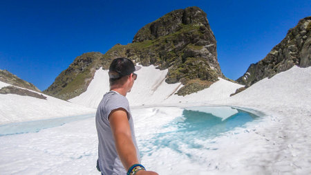 Young man talking a selfie with a frozen alpine lake. The ice shines in many different shades of...