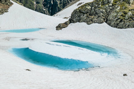 A view on a ice-frozen alpine lake. The ice shines in many different shades of blue. Lake is surrounded by tall mountains. There is no snow on the peaks. Spring slowly coming to high altitudesの写真素材